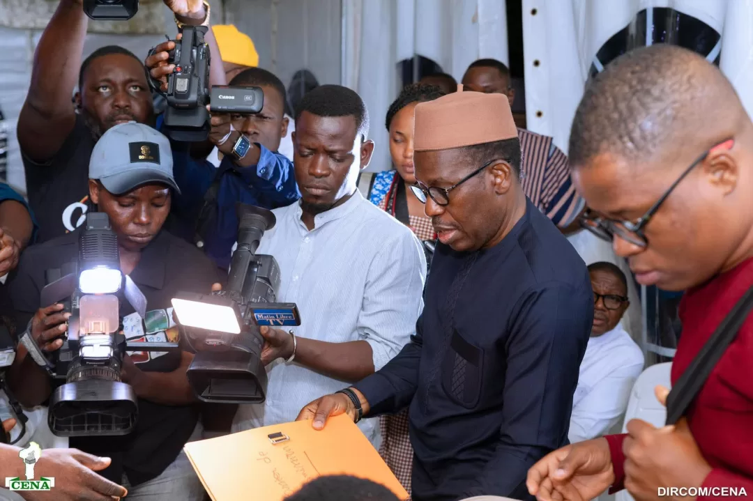 Un homme en tenue sombre tenant une enveloppe entoure de journalistes avec cameras et appareils photo allumes
