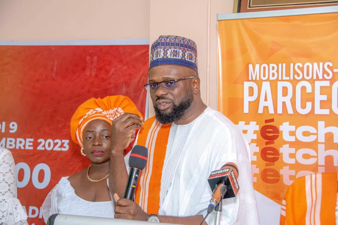 Un homme en tenue traditionnelle blanche et orange parle au micro devant un panneau orange tandis qu'une femme en tenue blanche et coiffe orange l'écoute.