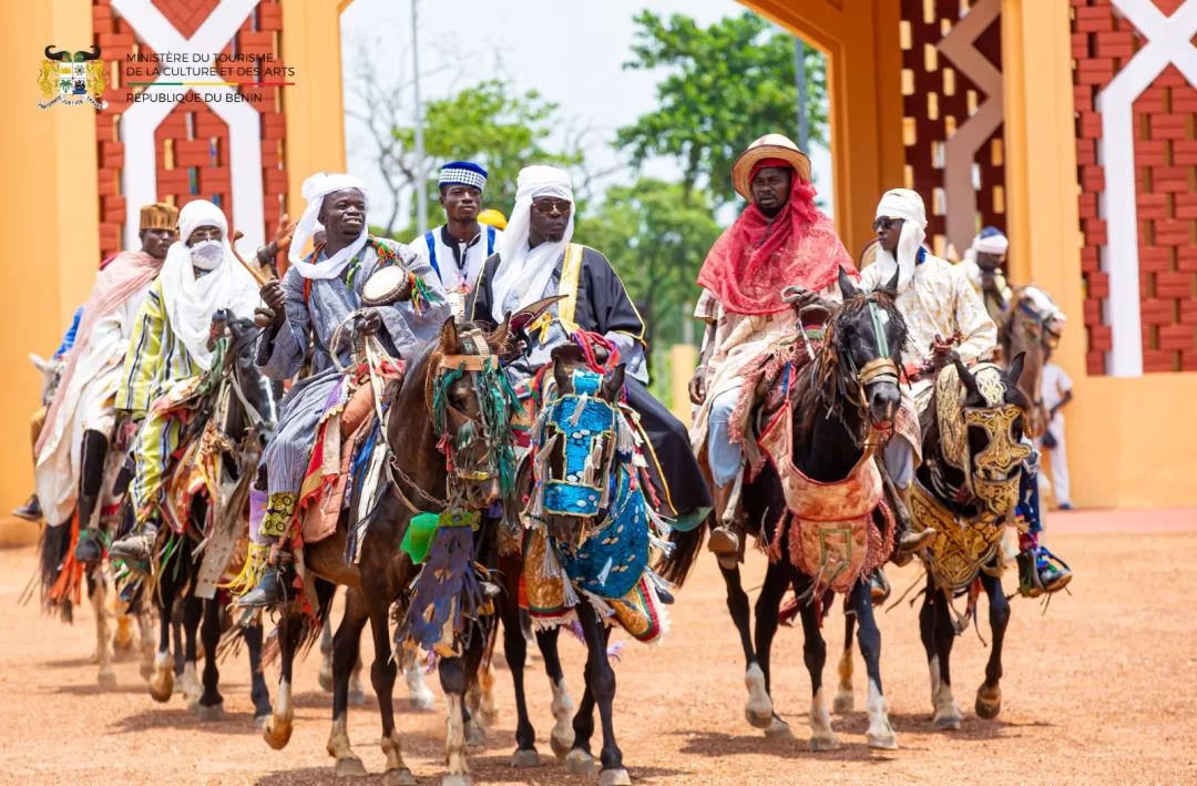 Un groupe d’hommes en habits traditionnels colorés montent des chevaux richement décorés au sol rouge devant un bâtiment aux murs ocres