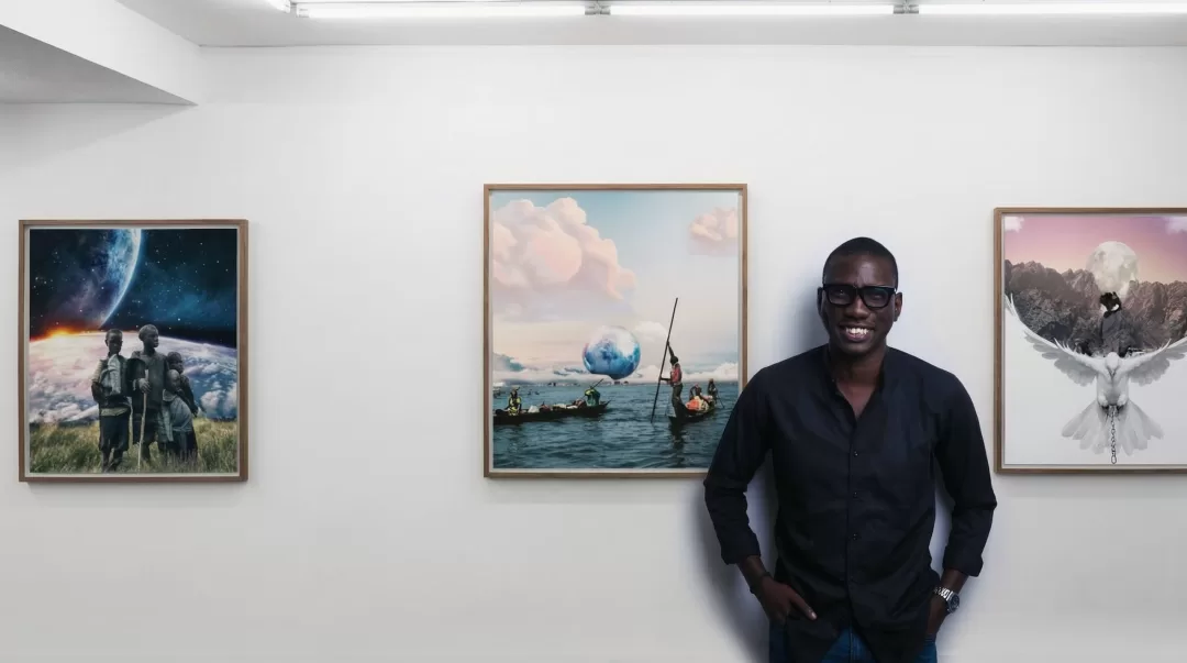 Un homme souriant en chemise noire pose devant trois tableaux accrochés dans une galerie d'art aux murs blancs