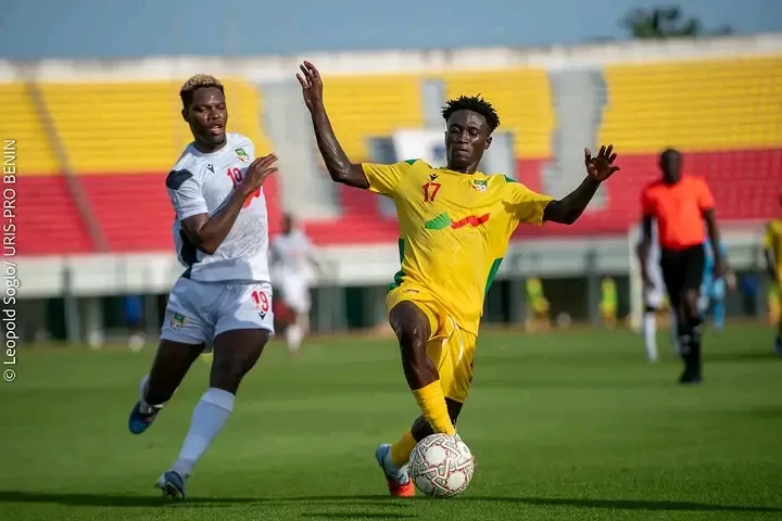 Deux joueurs de football en action : l’un en maillot jaune contrôle le ballon devant un adversaire en blanc dans un stade presque vide