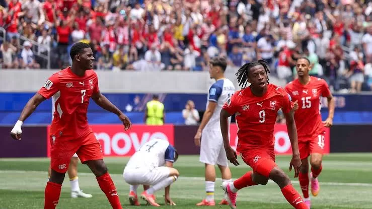 Des joueurs en maillots rouges célèbrent un but lors d’un match de football devant un public dense et des adversaires en blanc désabusés
