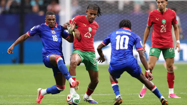 Deux joueurs en maillot rouge et deux en bleu bataillent pour le ballon au milieu d’un terrain de football