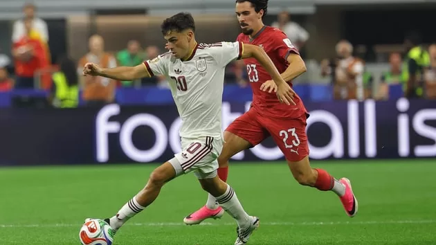 Deux joueurs de football en maillot blanc et rouge disputent le ballon sur le terrain sous le regard des spectateurs.