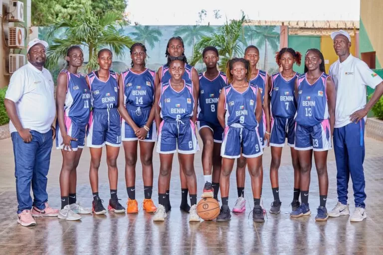 Groupe de jeunes basketteuses en tenue avec deux entraîneurs posant sur un terrain couvert avec un ballon de basket au centre