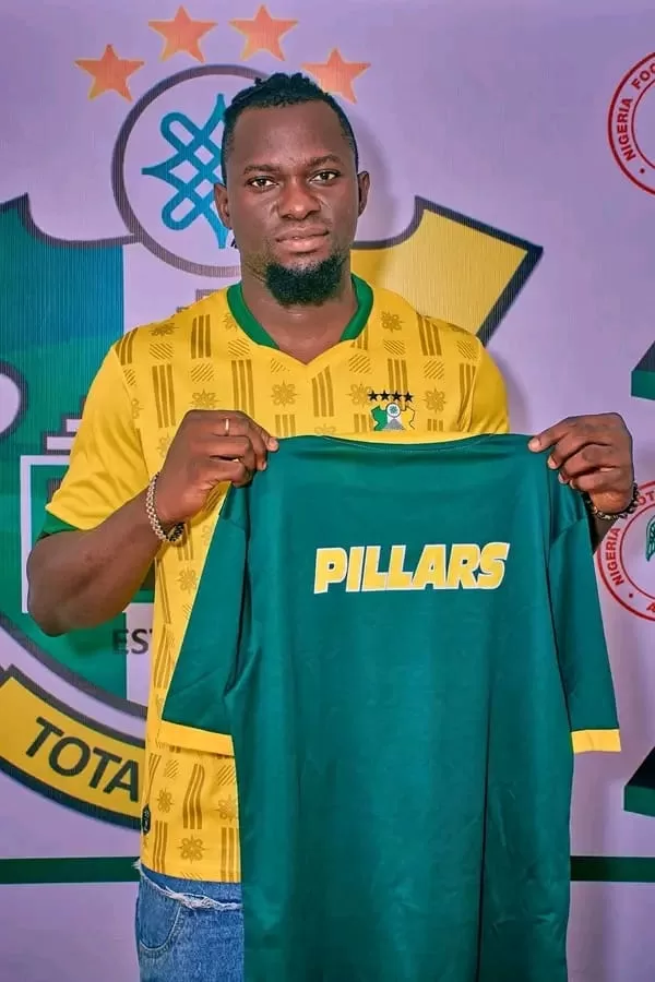 Un homme pose en tenue jaune tenant un maillot vert avec l'inscription PILLARS devant un logo sportif multicolore