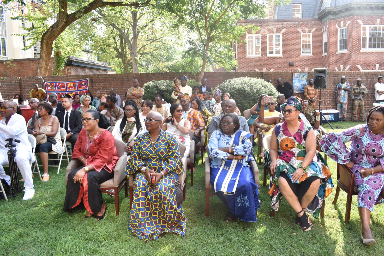 Groupe de personnes assises en plein air sur des chaises, certaines vêtues de tenues colorées africaines, devant un bâtiment en briques rouges