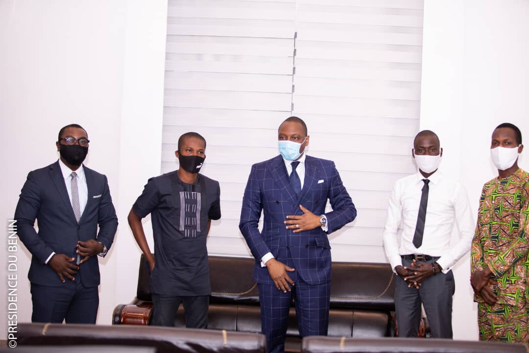 Cinq hommes debout devant un canapé, tous portant un masque et des vêtements formels ou traditionnels dans une pièce claire