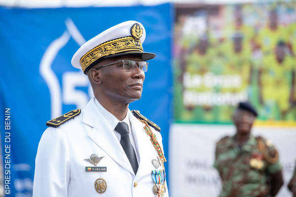 Un homme en uniforme blanc orné de médailles et décorations, portant une casquette et des lunettes, debout de profil