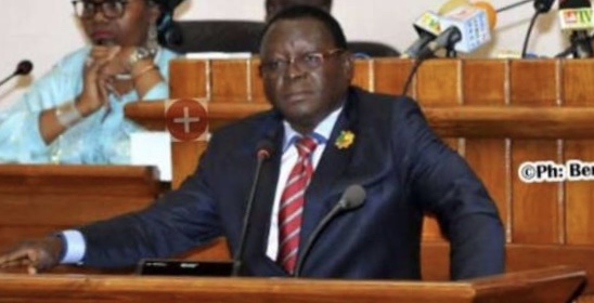 Un homme en costume sombre parle au micro dans un hémicycle avec une femme en arrière-plan.