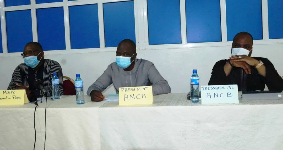 Trois hommes masqués assis à une table avec des étiquettes indiquant leurs fonctions dans une réunion formelle