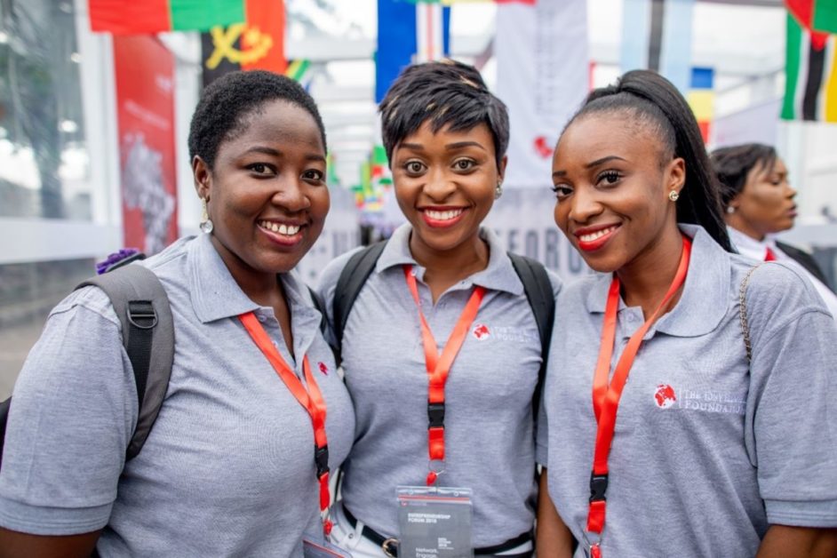 Trois jeunes femmes souriantes portant des polos gris et des badges rouges lors d’un événement en intérieur