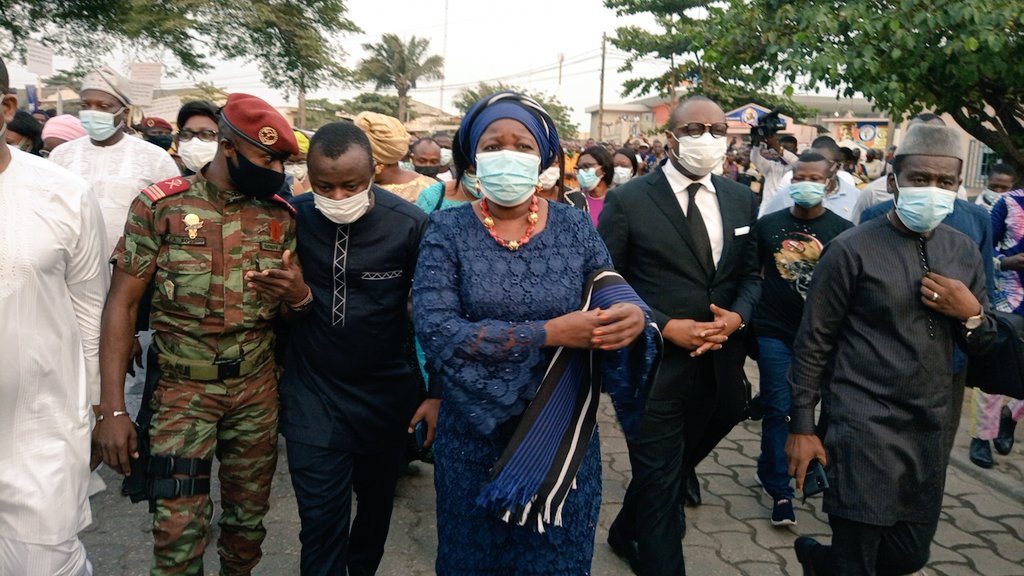 Un groupe de personnes masquées défile dans une rue, avec une femme en tenue traditionnelle bleue au centre entourée de plusieurs hommes dont un en uniforme mil