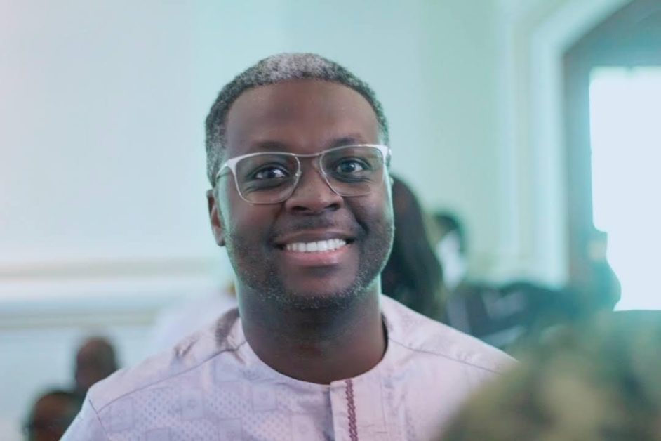 Homme souriant portant des lunettes et une tenue traditionnelle claire dans un intérieur lumineux et flou en arrière-plan