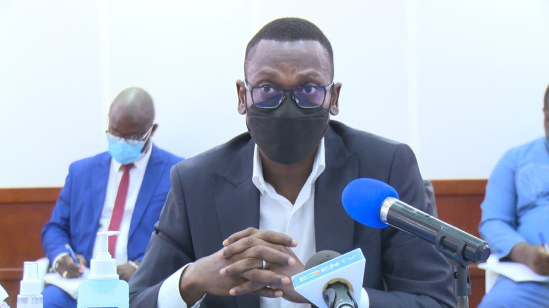 Un homme en costume sombre et lunettes porte un masque noir, assis derrière un micro bleu lors d'une réunion avec d'autres personnes.