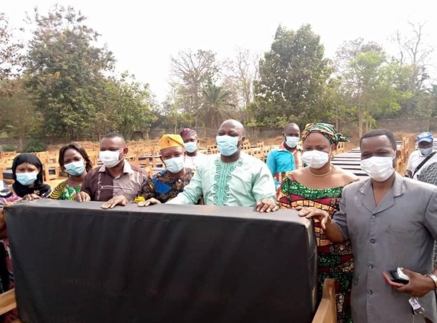 Un groupe de personnes masquées tenant un lit avec matelas devant de nombreux autres lits alignés en extérieur avec des arbres en arrière-plan