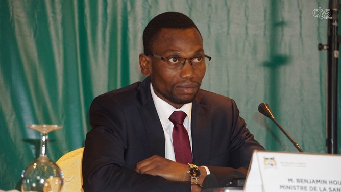 Homme en costume sombre et cravate assis à une table avec microphone face à un rideau vert.