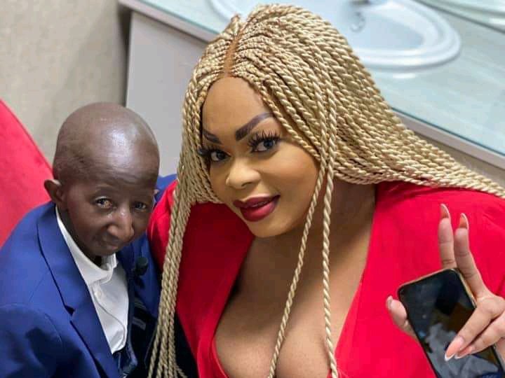 Un homme en costume bleu et une femme en robe rouge avec de longues tresses blondes posent ensemble, elle fait un signe de paix avec la main