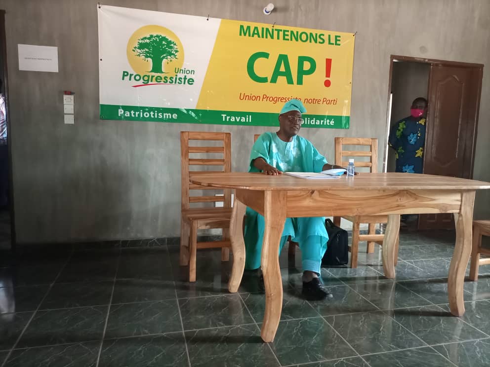Un homme en tenue traditionnelle bleue est assis derrière une grande table en bois devant une banderole jaune et verte sur un mur gris