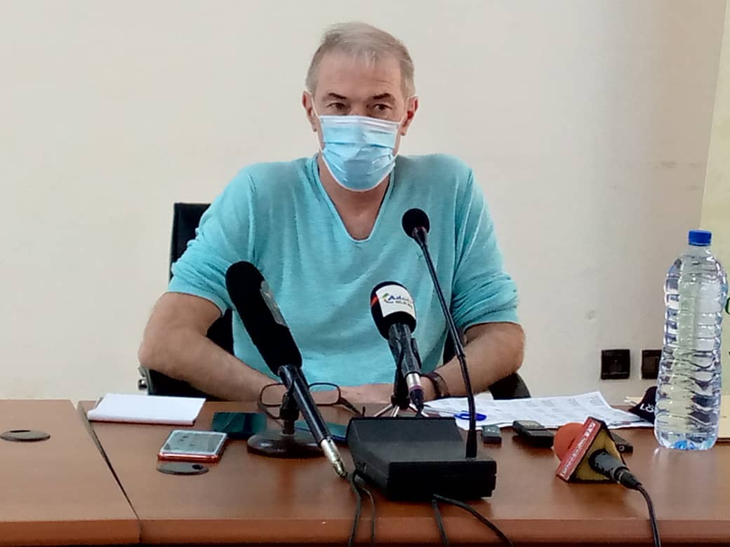 Homme masqué en chemise bleue assis derrière une table avec plusieurs micros et une grande bouteille d'eau