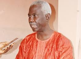 Homme âgé en tenue traditionnelle rouge parlant face à un microphone tenu par une main extérieure