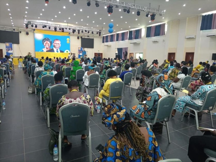 Des dizaines de femmes assises dans une grande salle écoutent des intervenants sur une scène avec un grand écran coloré
