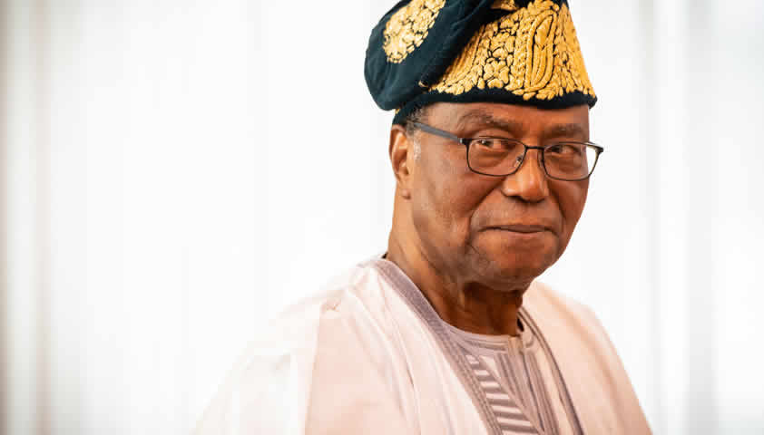 Homme portant une tenue traditionnelle claire et un chapeau orné souriant légèrement devant un fond blanc lumineux