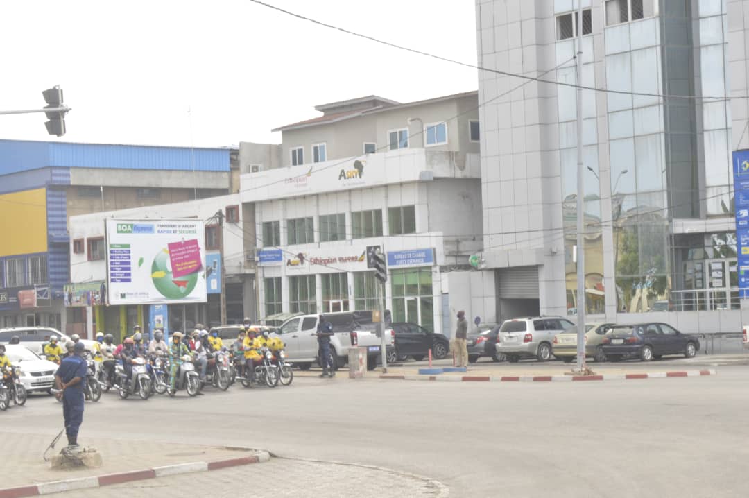 Un groupe de motards et plusieurs piétons se tiennent à un feu tricolore dans un carrefour urbain devant des bâtiments modernes