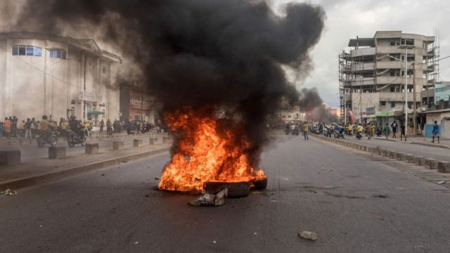Un feu intense dégage une épaisse fumée noire au milieu d’une rue urbaine avec des bâtiments et des passants en arrière-plan
