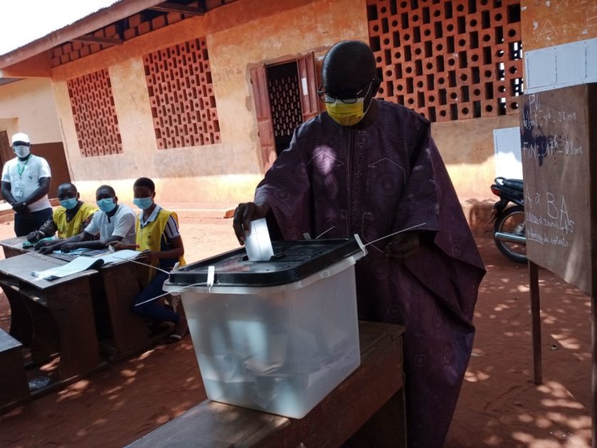 Un homme masqué dépose un bulletin dans une urne transparente sur fond de bâtiment et de personnes assises au bureau de vote