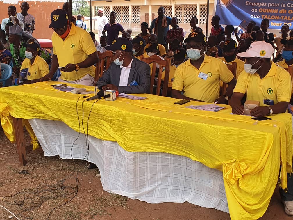 Personnes assises à une table couverte de tissu jaune et blanc, portant des polos jaunes et casquettes noires avec logo, en extérieur sous abri