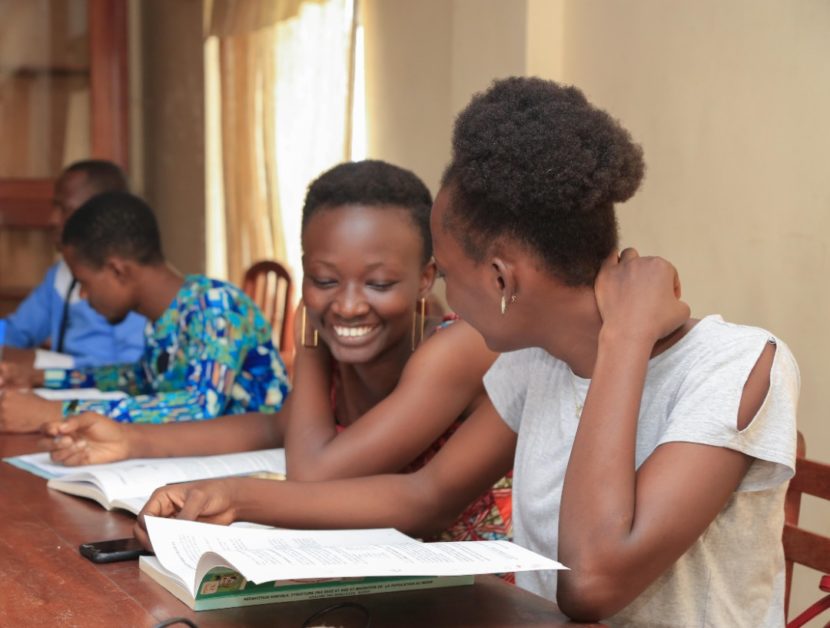 Deux femmes souriantes lisent un livre ensemble assises à une table dans une salle avec d’autres personnes en arrière-plan