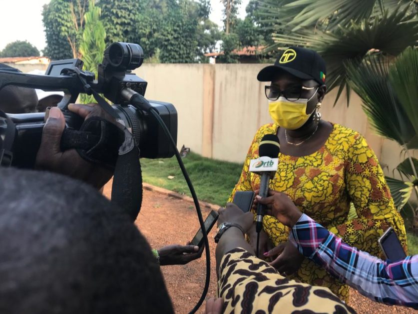 Une femme en tenue jaune et masque parle face à plusieurs journalistes armés de micros et caméras dans un espace extérieur vert