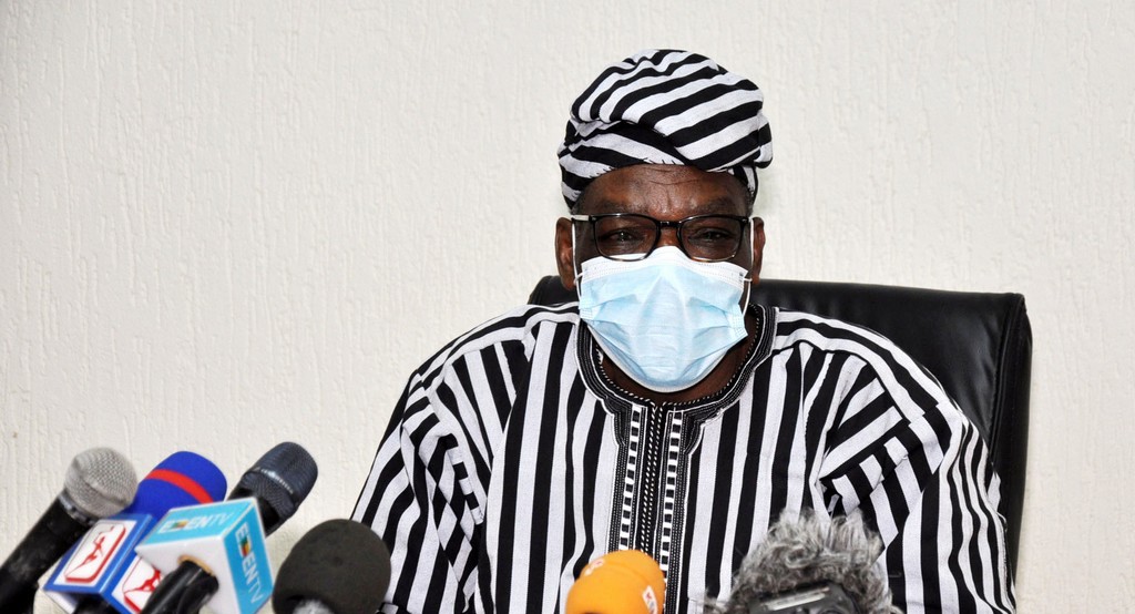 Homme en pagne rayé noir et blanc avec masque et lunettes assis derrière plusieurs micros sur table devant un mur blanc
