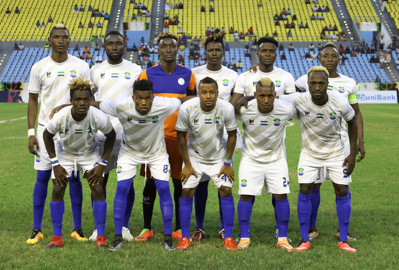 Onze joueurs de football en tenue blanche posent sur un terrain avant un match dans un stade avec des gradins jaunes et bleus