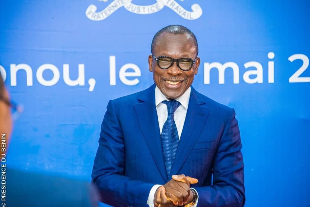 Un homme en costume bleu souriant debout devant un mur bleu avec des inscriptions blanches partiellement visibles