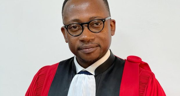 Homme portant une robe judiciaire rouge et noire avec une cravate blanche et des lunettes noires regarde l’objectif avec un léger sourire