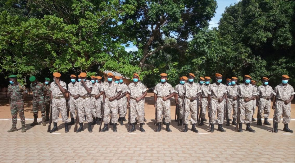 Un groupe de militaires en uniforme clair et portant des masques, alignés en rangs dans un espace extérieur arboré
