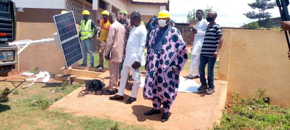 Un groupe d hommes, certains en tenue traditionnelle et d autres avec casque jaune, examine un panneau solaire posé au sol devant un bâtiment beige