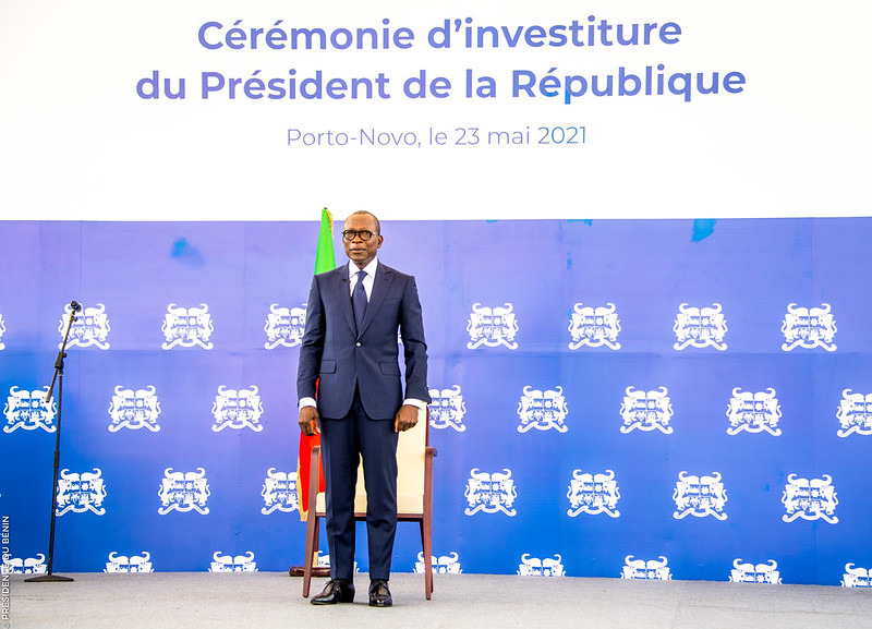 Un homme en costume sombre se tient debout devant un fond bleu avec texte et armoiries blanches et un drapeau visible derrière lui
