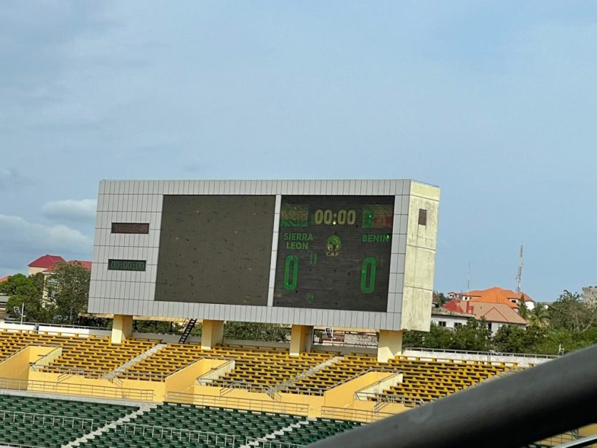 Un tableau d'affichage dans un stade indique 0-0 et le chronomètre à 00:00 au-dessus des gradins jaunes et verts