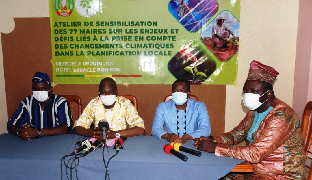 Quatre hommes portant des masques sont assis derrière une table avec des microphones devant une affiche sur un atelier de sensibilisation aux changements climat