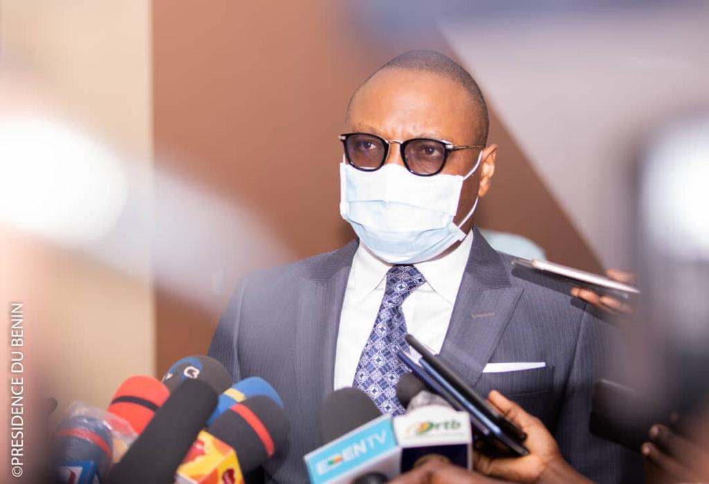 Homme en costume, lunettes et masque blanc parlant devant plusieurs micros de médias