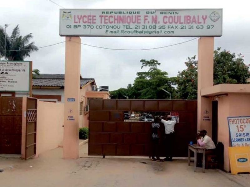 Entrée d un lycée technique avec portail fermé et deux personnes discutant à côté d une table sur la droite
