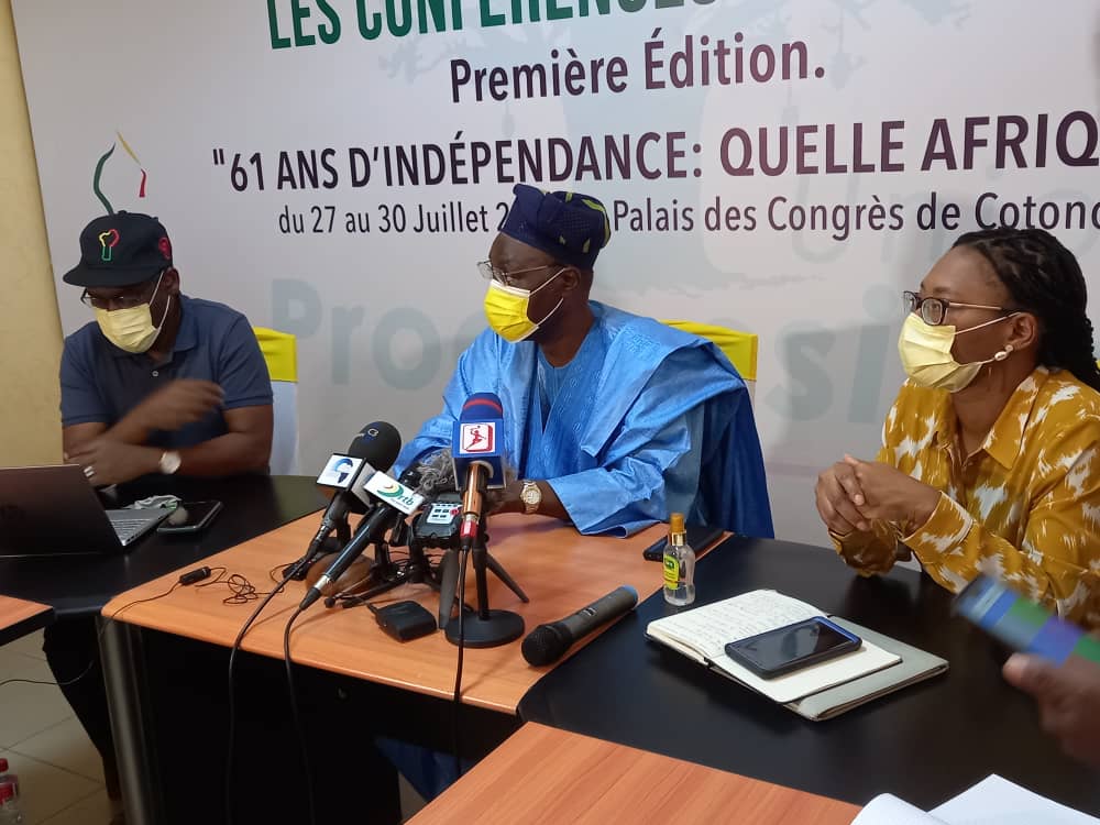 Trois personnes masquées assises autour d'une table avec microphones et matériel de prise de notes devant un panneau d'exposition textuel