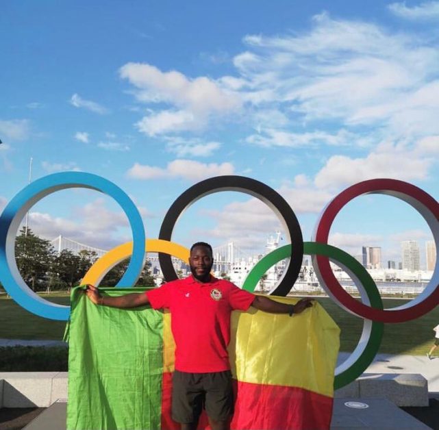 Un homme en tenue sportive rouge tient un drapeau tricolore devant les anneaux olympiques en extérieur