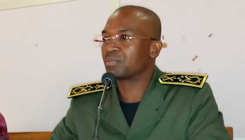 Homme en uniforme militaire vert parlant devant un microphone dans une salle au mur blanc