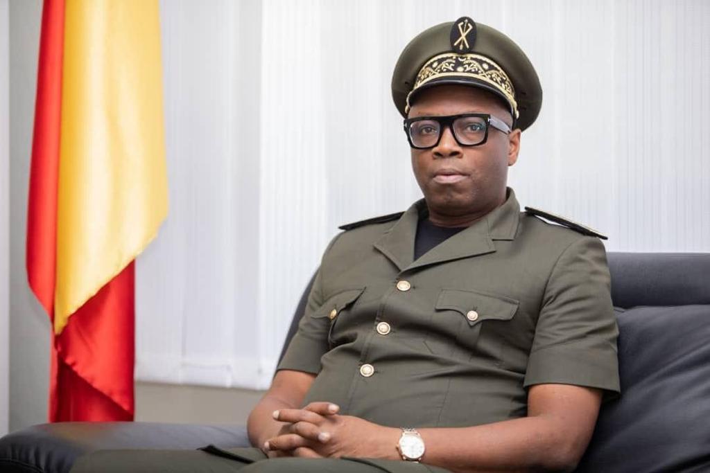 Homme en uniforme militaire assis sur un canapé devant un drapeau aux couleurs rouge, jaune et vert