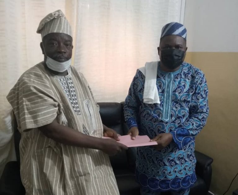 Deux hommes en tenue traditionnelle et masque tenant un document rose dans une pièce avec un canapé noir et un rideau beige