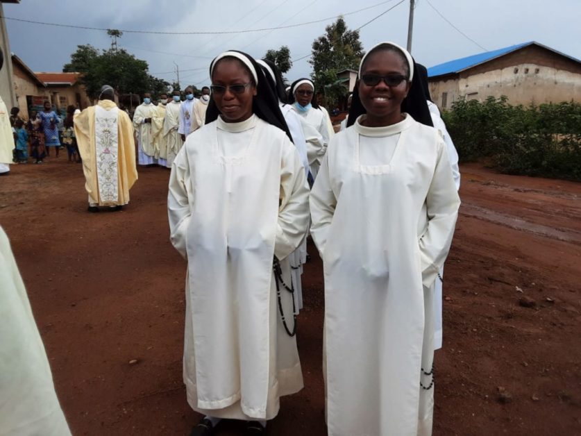 Deux religieuses en habits blancs marchent sur une route de terre rouge devant un groupe de personnes en arrière-plan